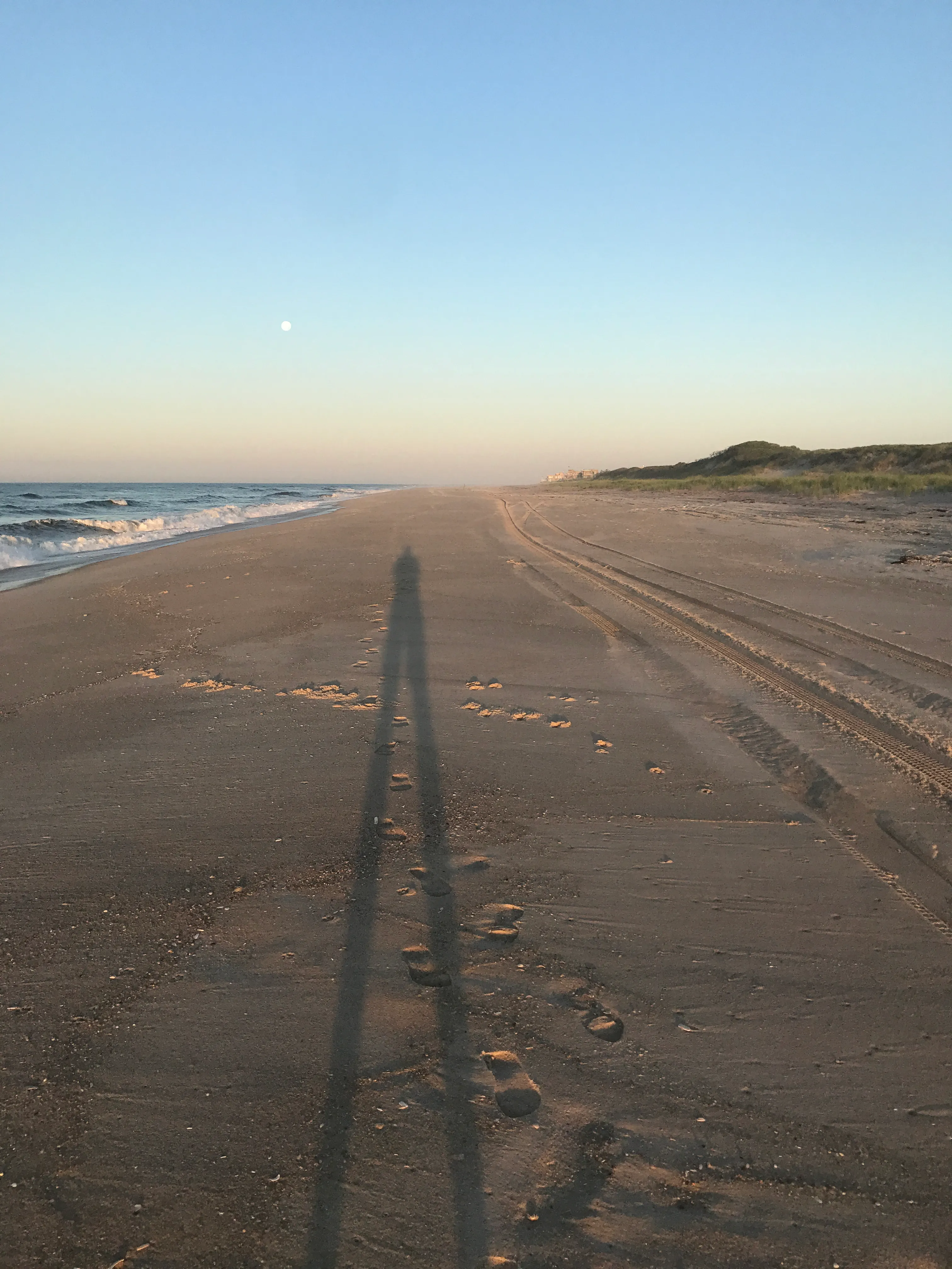 Long shadow of a person cast across wet sand at low tide, Fire Island shoreline at golden hour.