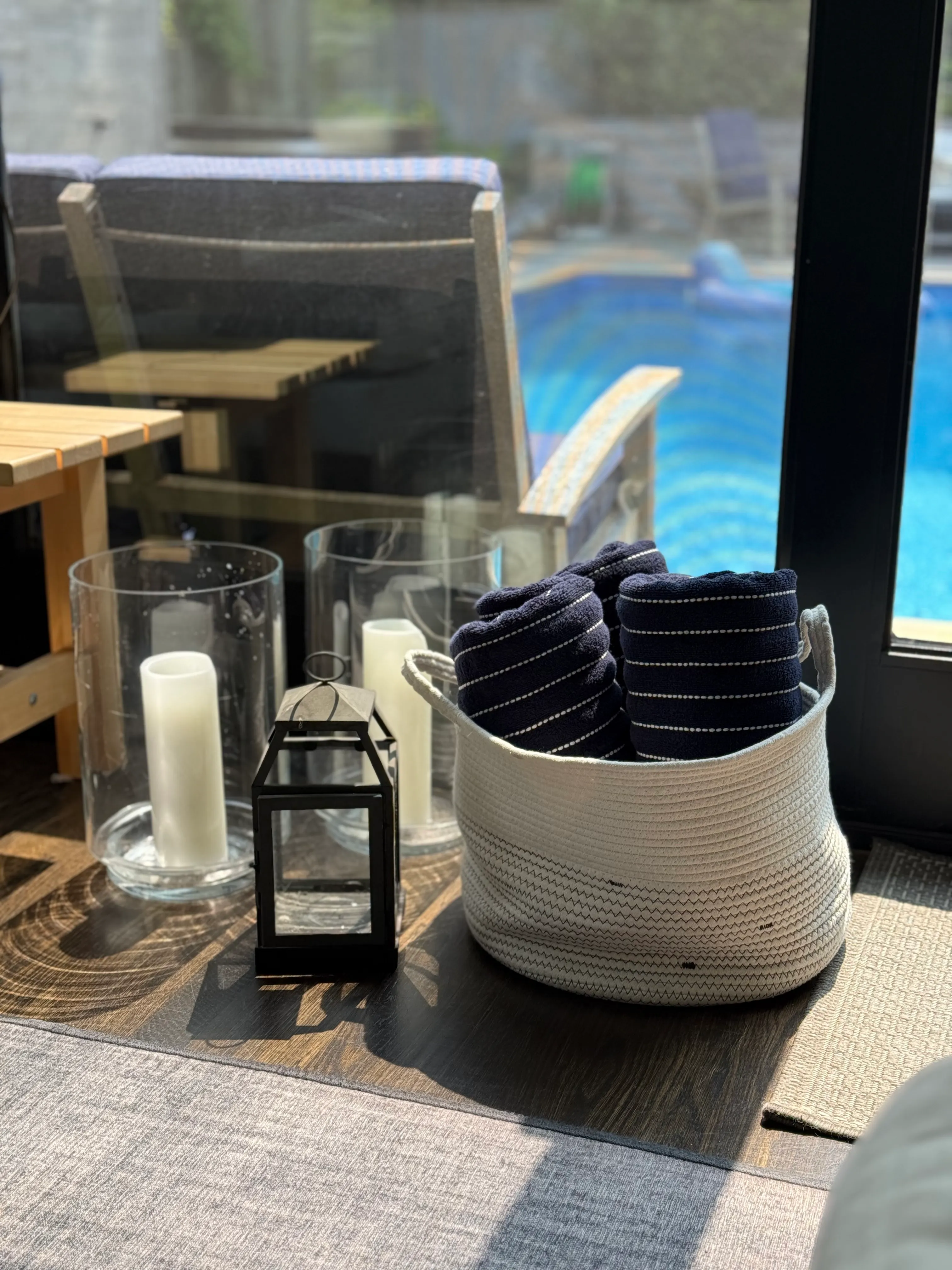 Poolside patio with a basket of rolled navy towels, lit candles in hurricane lamps, and a teak chair.