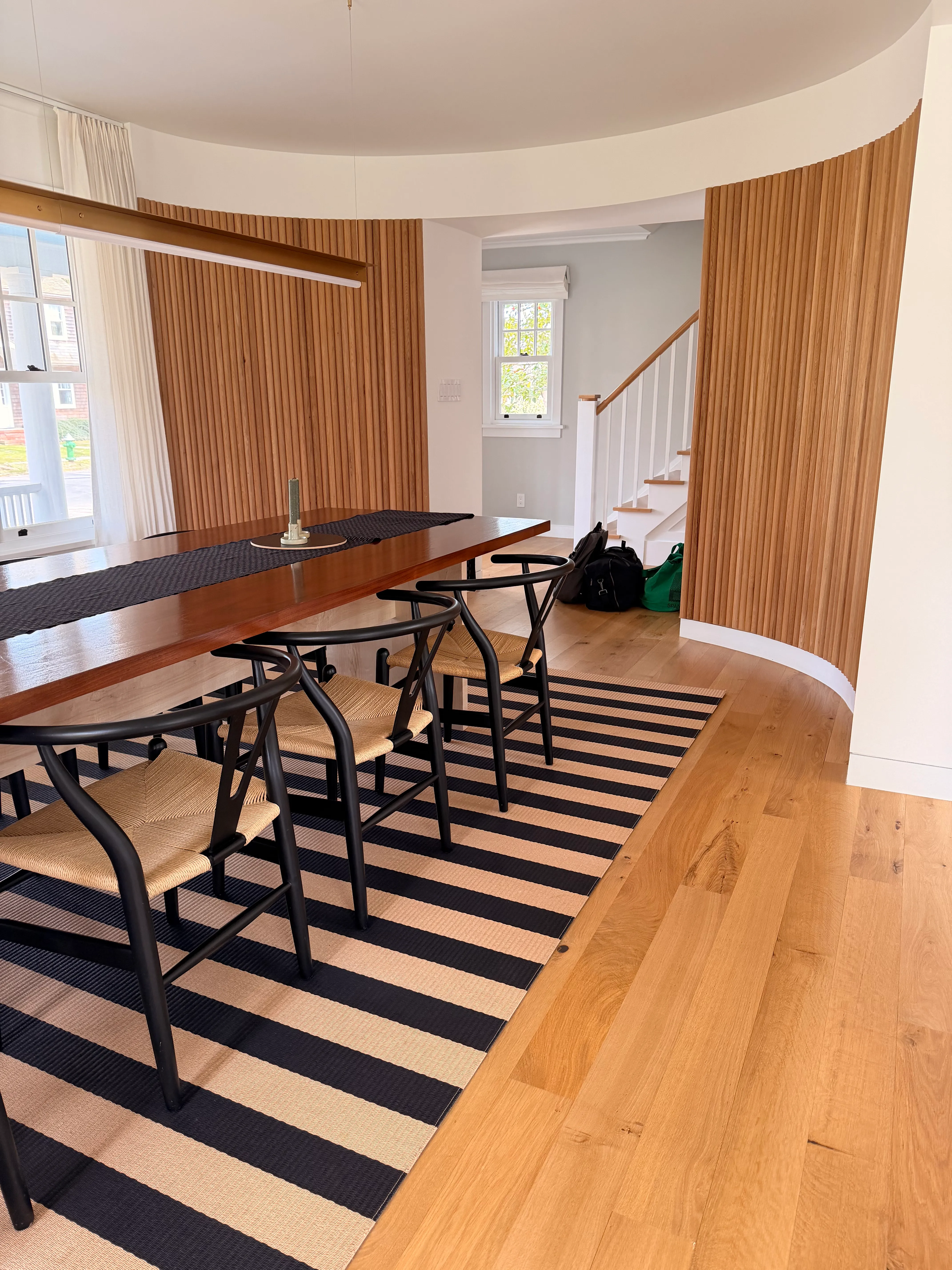 A dining room with black wishbone chairs, a striped rug, and a curved wall of pale wood slats.