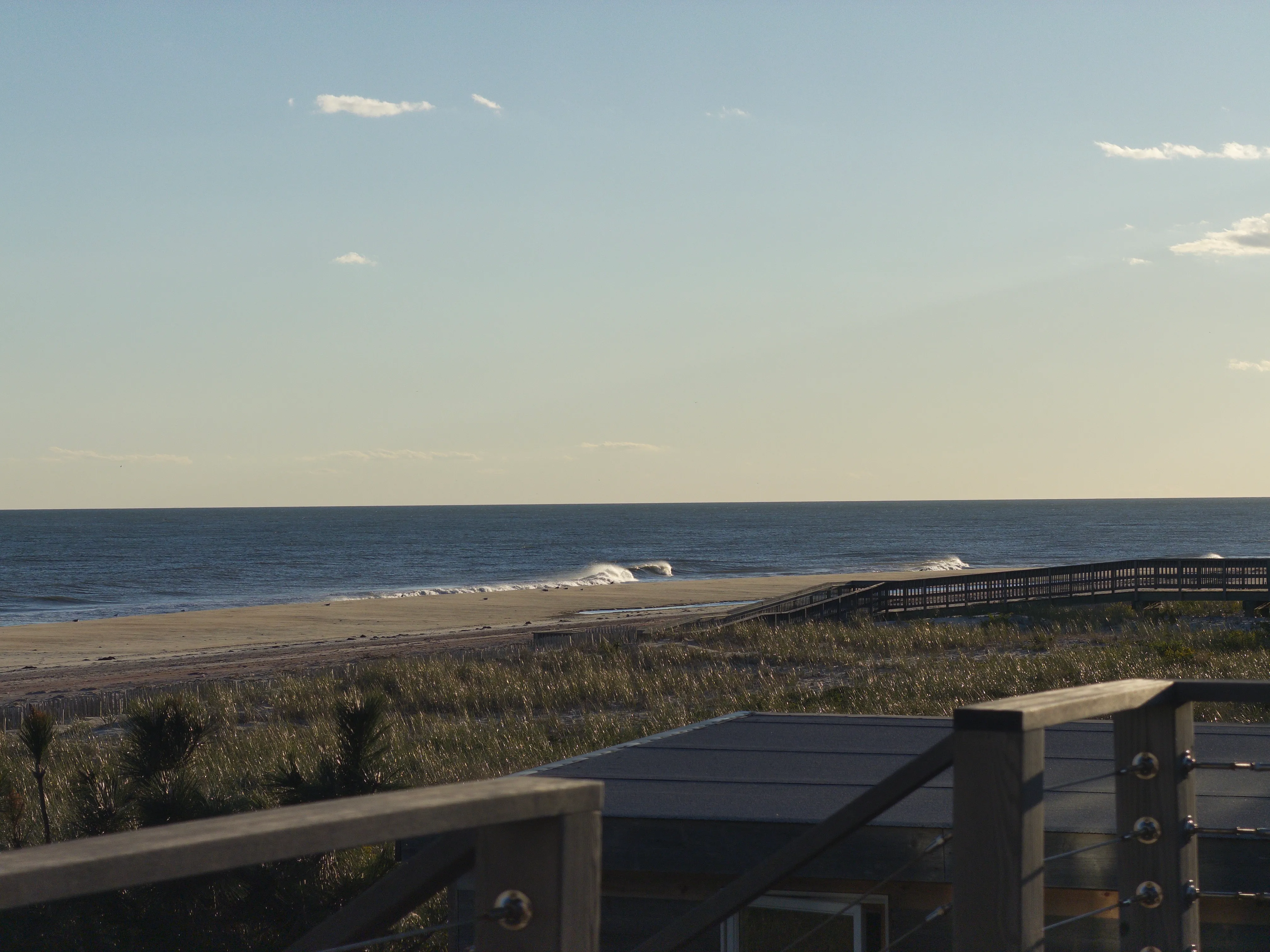 The view from a Fire Island Pines deck: cable railing, dune grass, a boardwalk to the beach, and surf breaking in late-afternoon light.