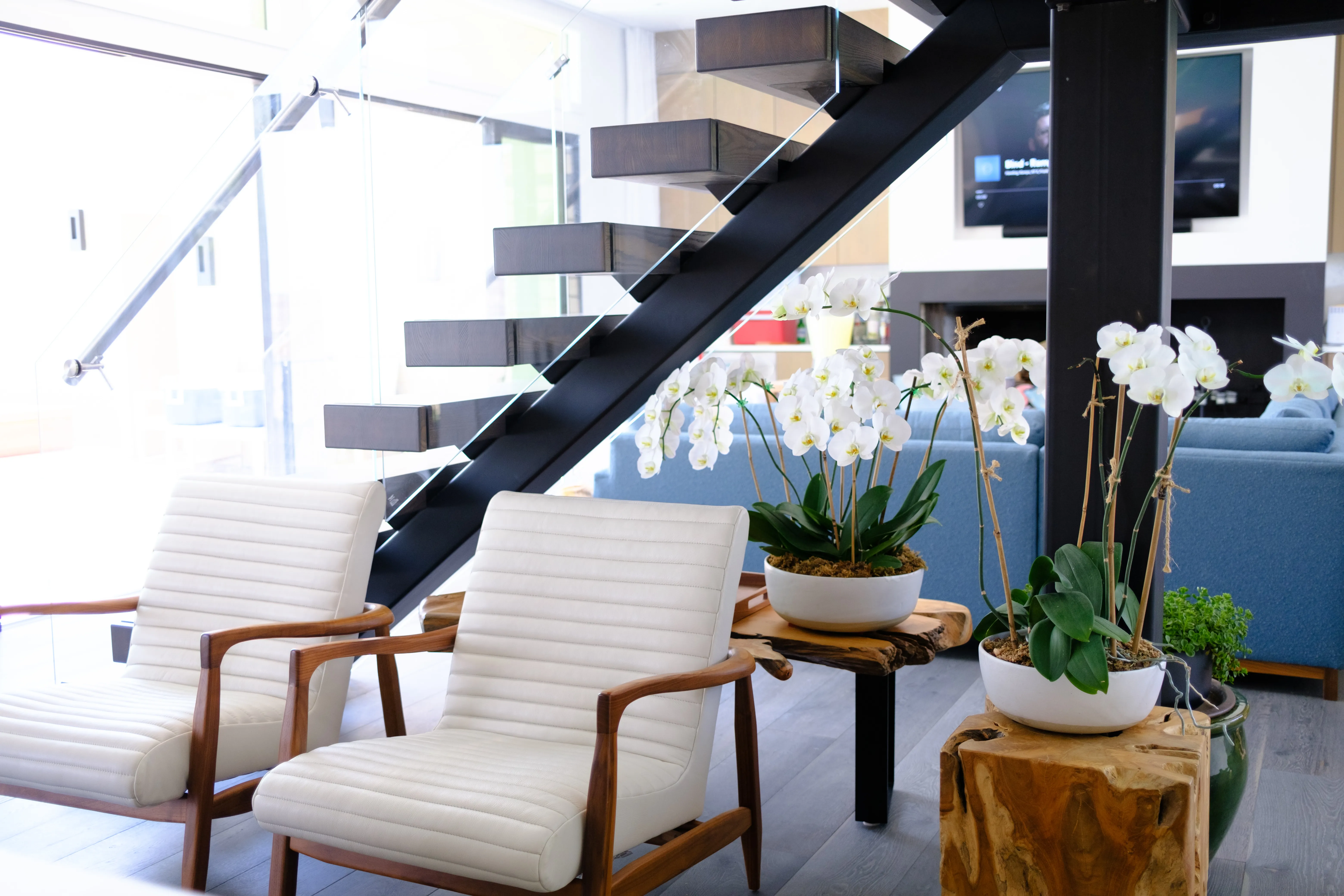 A modern entry with white lounge chairs, a black floating staircase, and orchids on tree-stump tables — the house ready for a weekend.