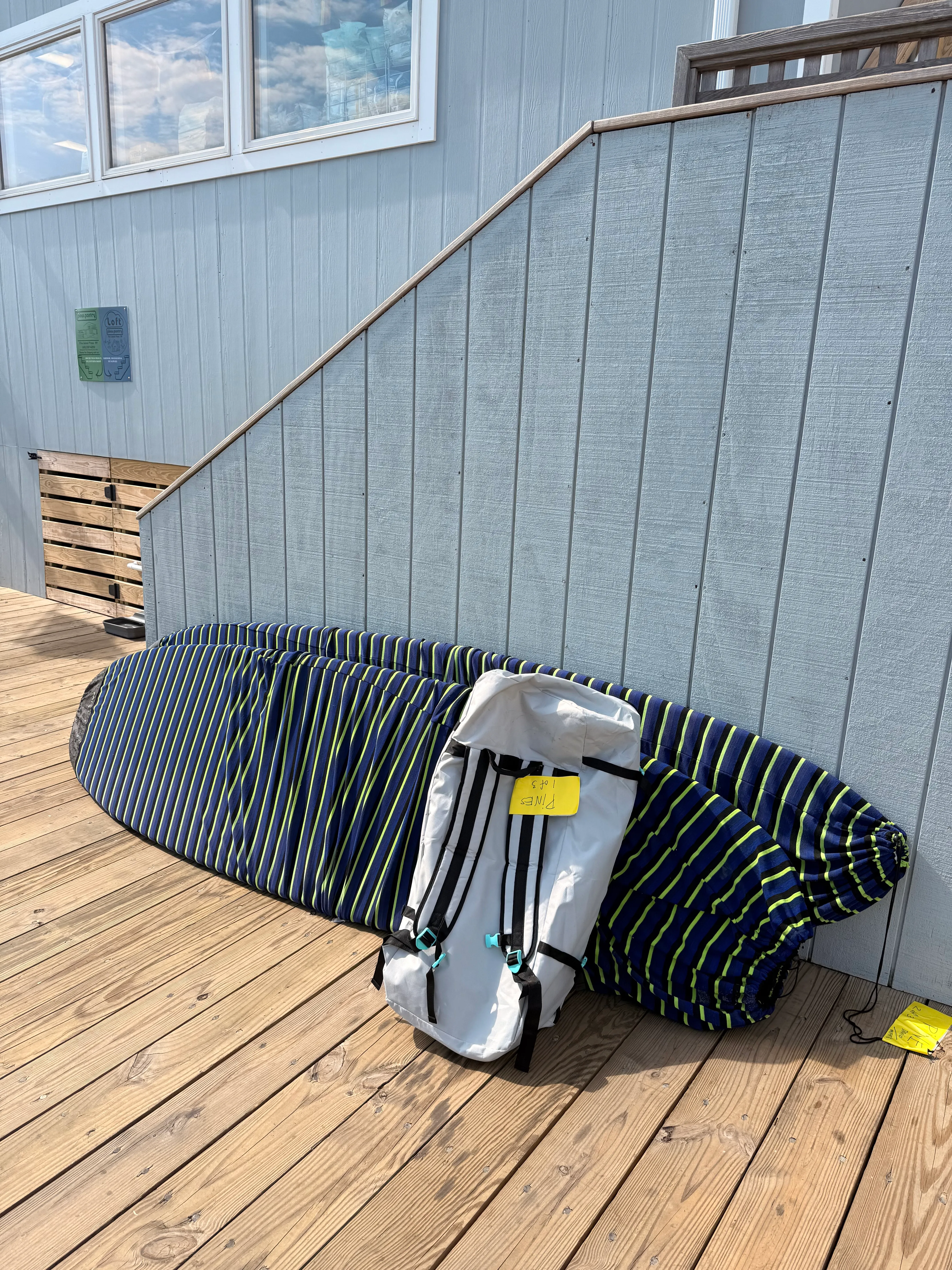 Two stand-up paddleboards in navy and lime striped board socks, with a white gear bag tagged for the Pines, leaning against grey siding on a wooden deck.