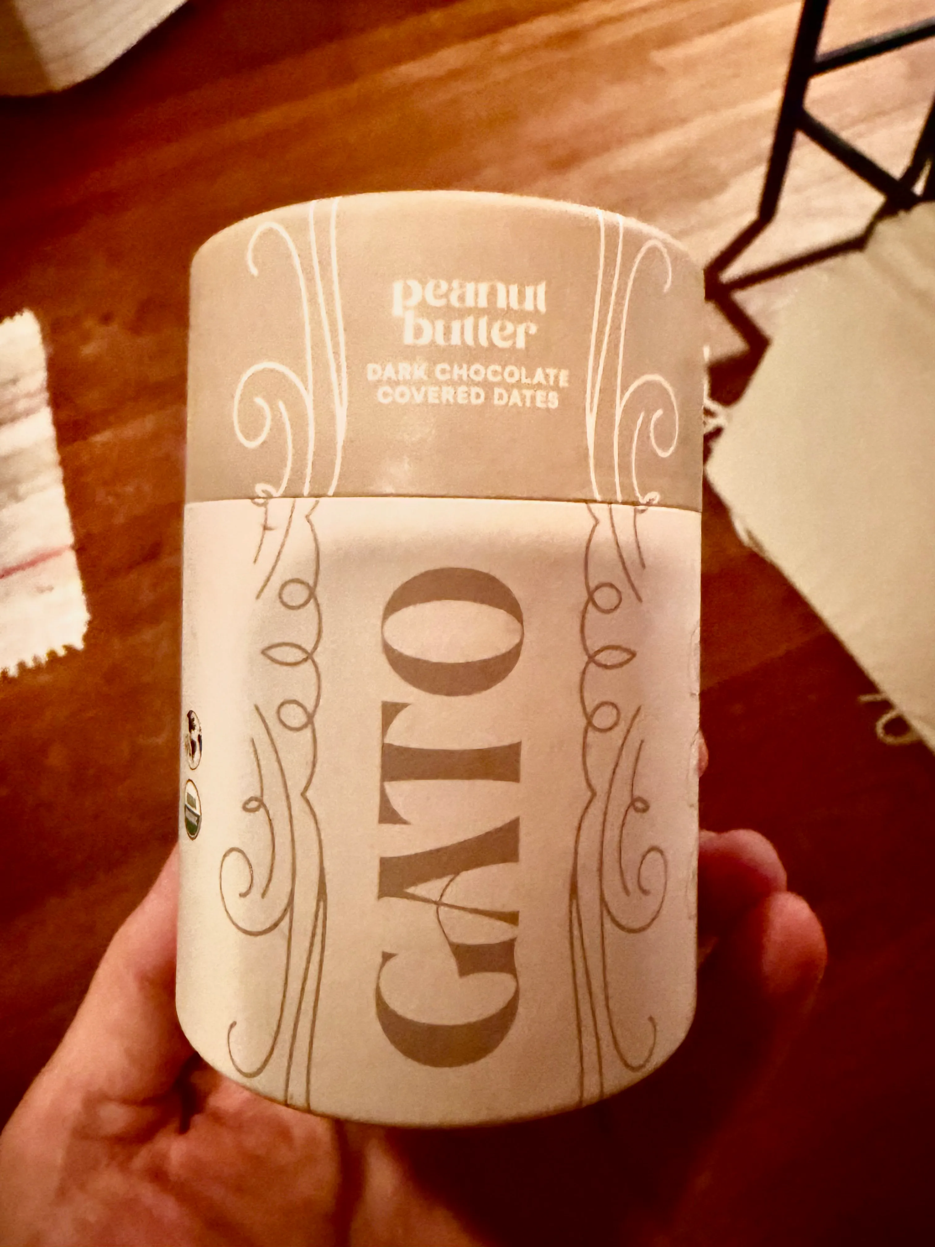 A hand holding a cream-colored GATO canister of peanut butter dark chocolate covered dates.